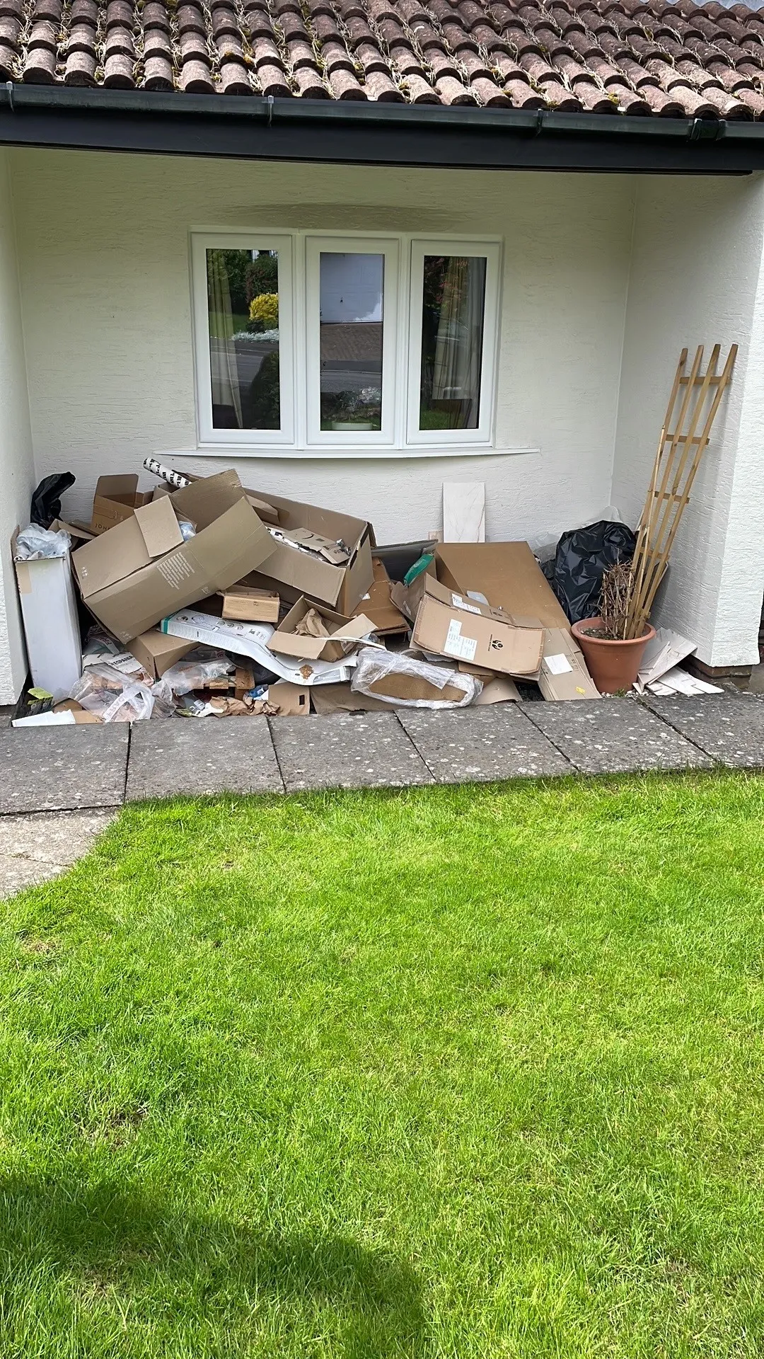 A pile of cardboard boxes, packaging materials, and rubbish is stacked under a house window near a green lawn—ideal for quick Rubbish Clearance. A plant pot and wooden trellis sit on the right side.