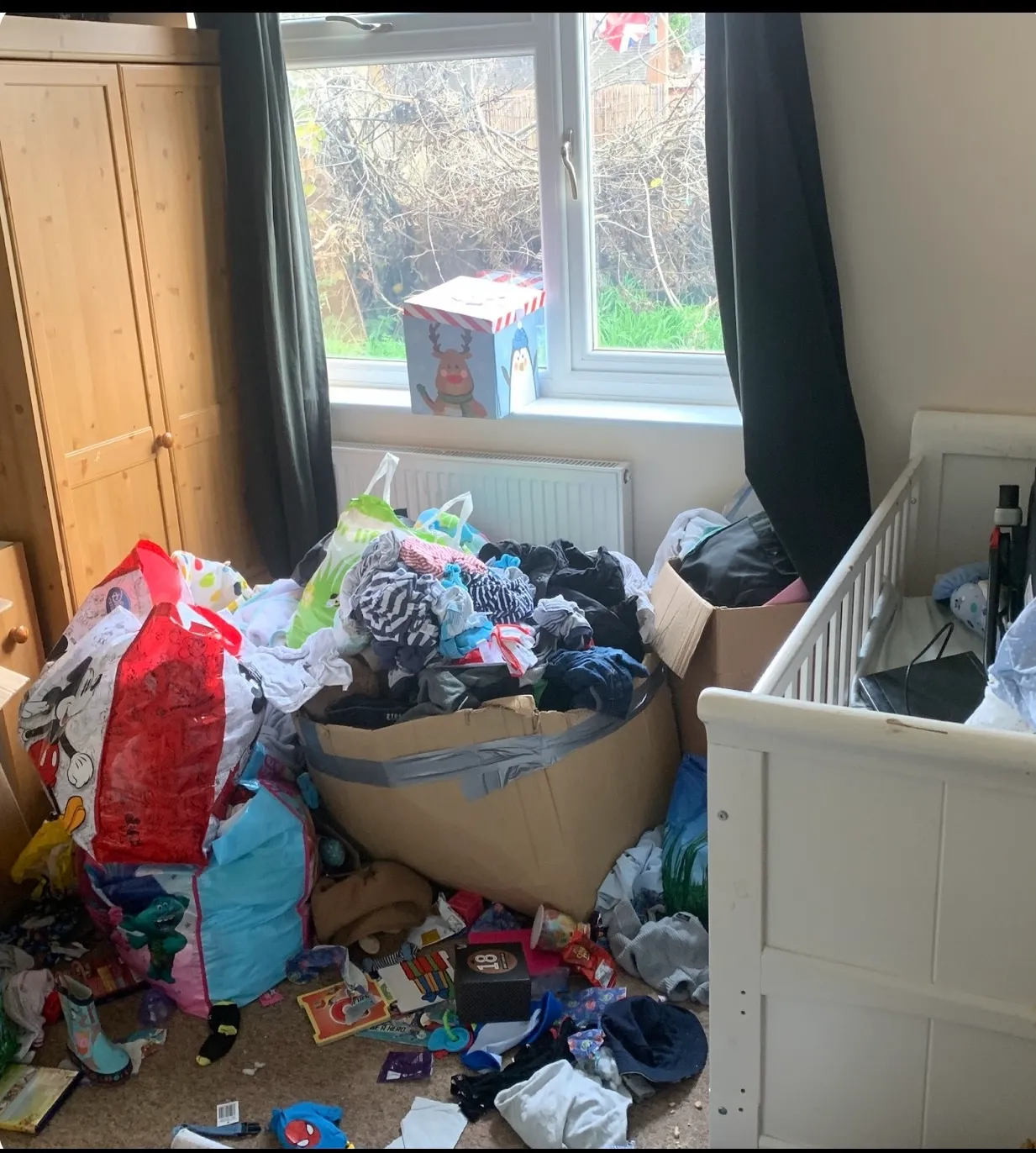 A cluttered bedroom with piles of clothes, toys, and bags on the floor next to a crib and wardrobe. Light seeps through black curtains, highlighting a space in need of house clearance to restore order and cleanliness.