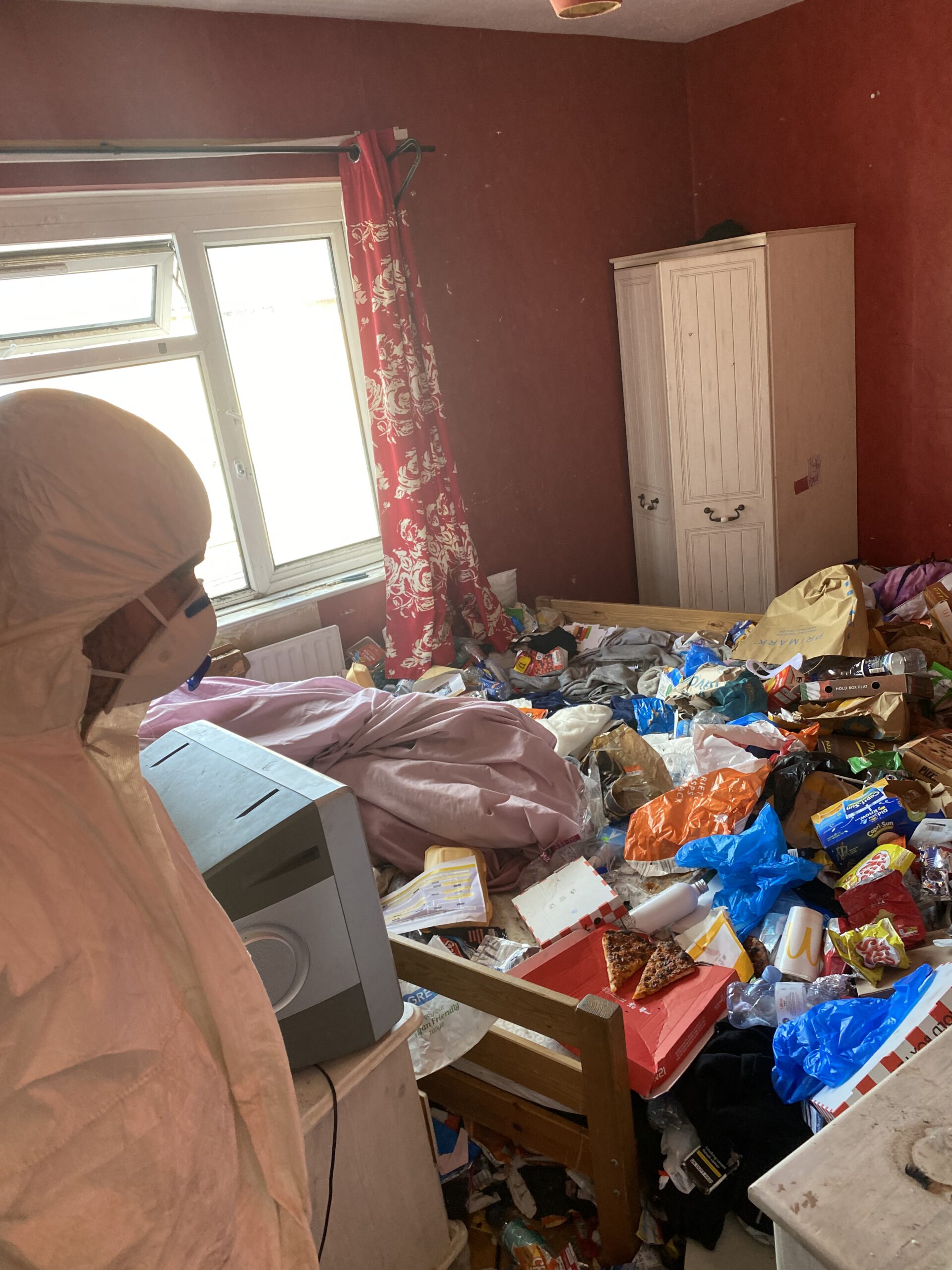 A person in protective clothing and a face mask stands in a cluttered room filled with trash and scattered items, sunlight streaming through an open window with red floral curtains—ideal for a House Clearance (+5 Variables See Questionnaire) service.