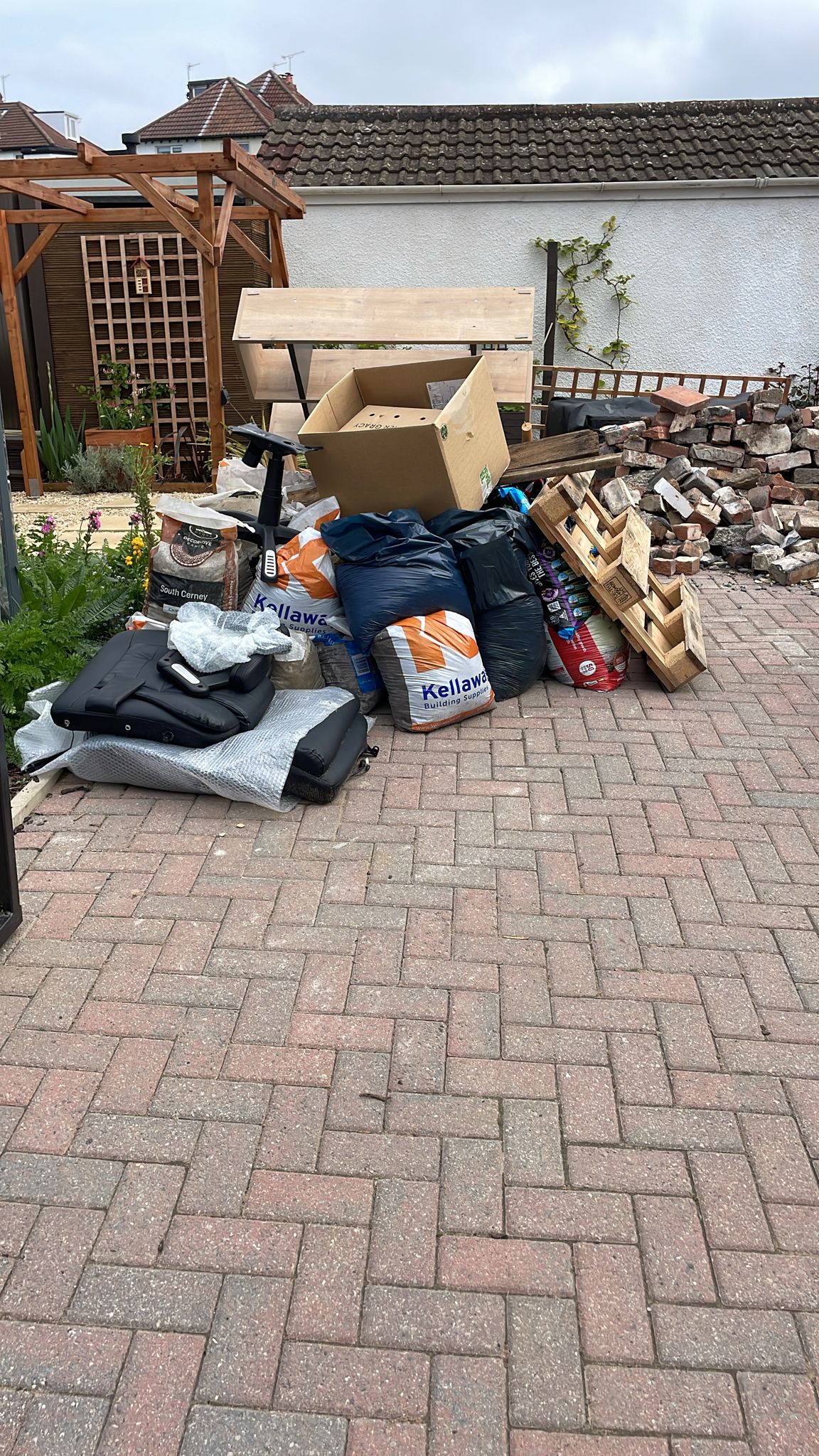 A pile of assorted waste sits on a paved driveway next to a garden with a wooden trellis and white wall, perfect for a quick and efficient Rubbish Clearance or House Clearance service.
