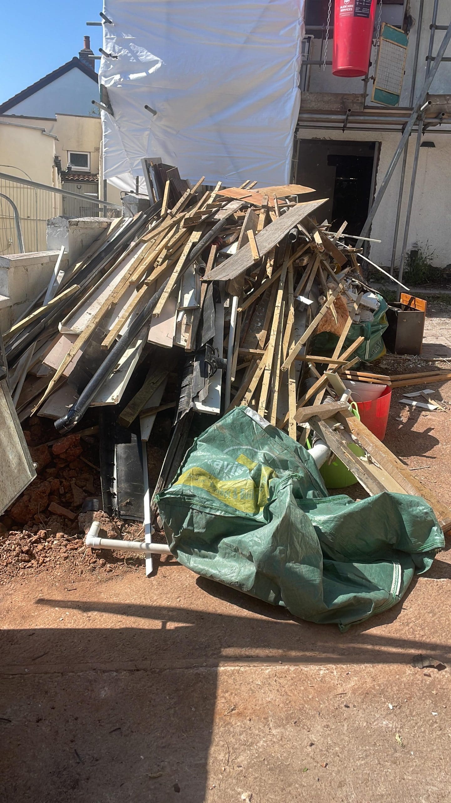 A large pile of construction debris, including wooden planks and metal pieces, sits on the ground at a building site, ready for professional Site Clearance. Green tarps and building materials are scattered nearby, with a partially constructed building in the background.