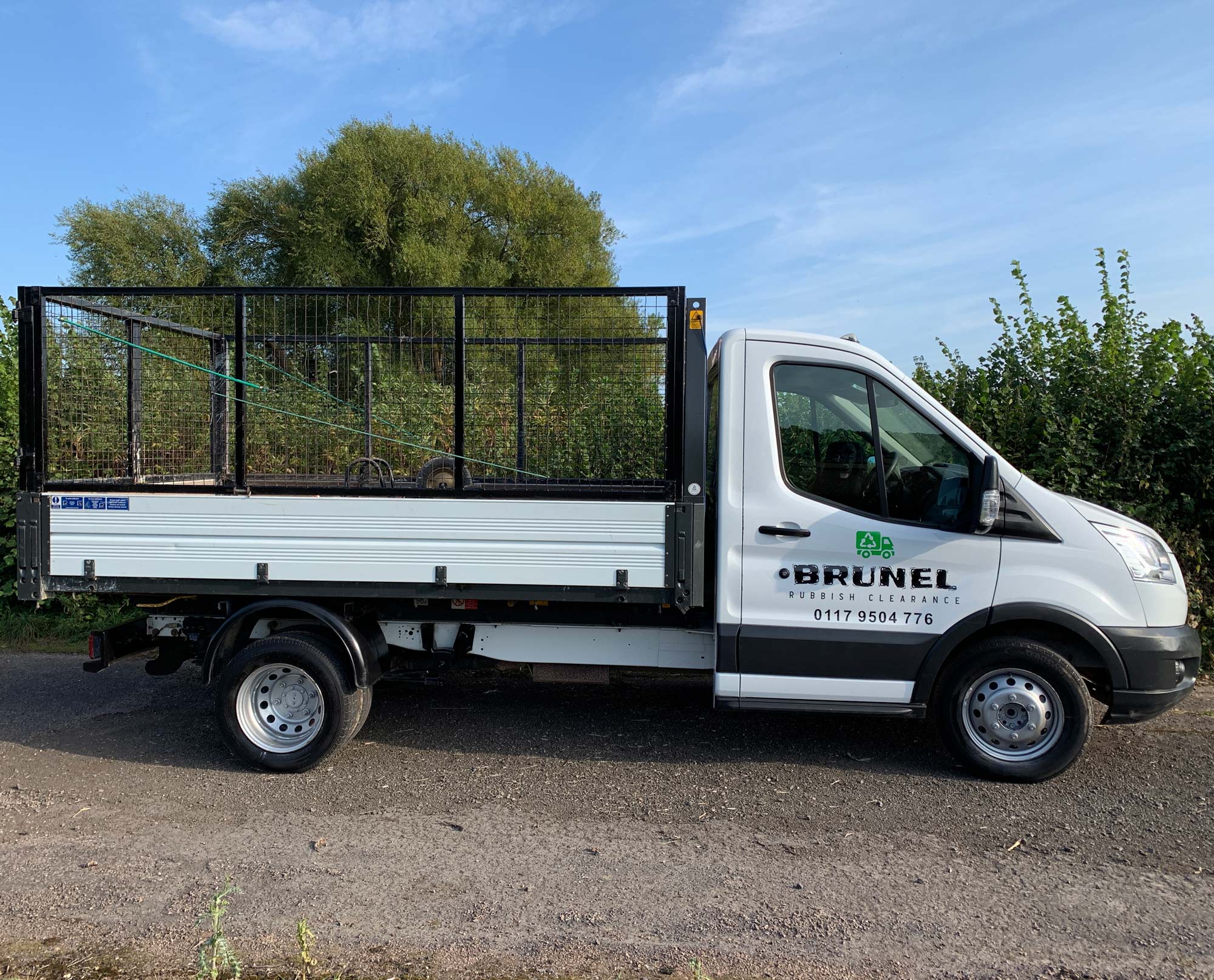 A white Brunel Rubbish Clearance truck with a metal cage on its flatbed is parked on a roadside beside green bushes and trees, under a clear blue sky.