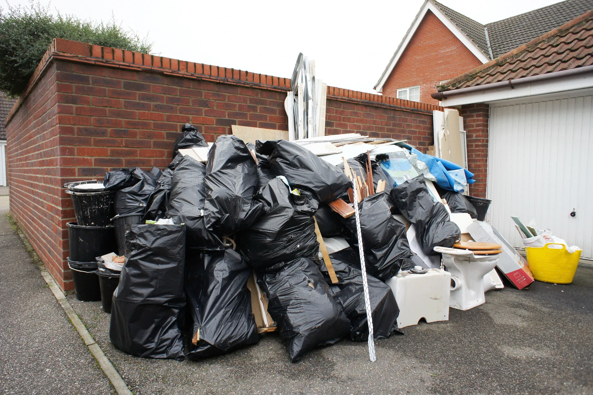 A large pile of black garbage bags and assorted rubbish, including household items and debris, awaits House Clearance next to a garage against a brick wall in a residential area.