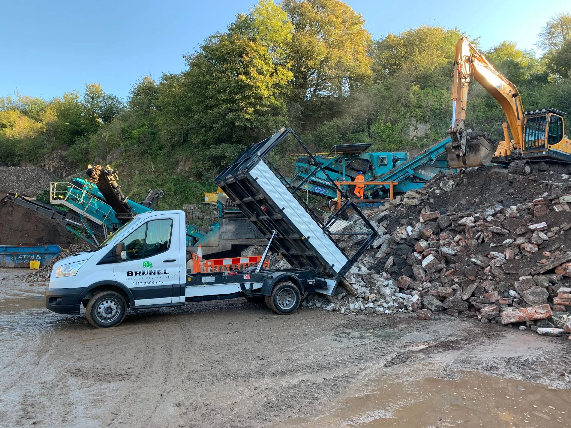 A white tipper truck unloads bricks and rubble at a construction site during site clearance, while an excavator and workers operate nearby amidst piles of debris and trees in the background.